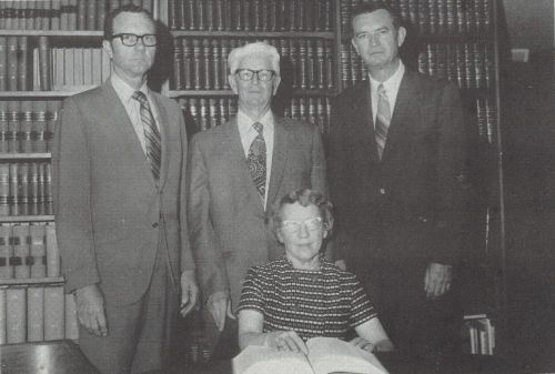 Image of the first State Law Library board, composed of four members. Three members are pictured standing and the fourth sitting in front of a bookcase of legal volumes.