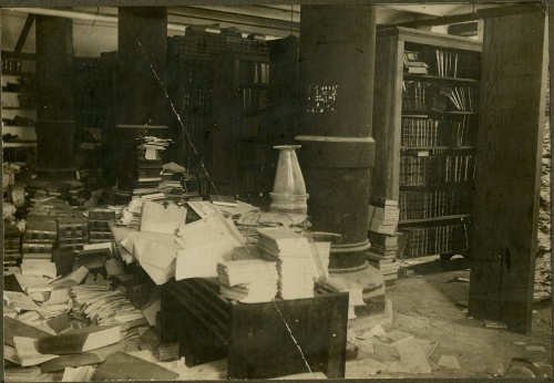 Image of the Supreme Court Library in the basement of the Capitol, ca. 1909, a basement room with overflowing shelves and leaning piles of books stacked haphazardly on desks and the floor.