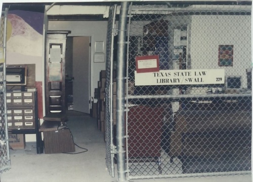 Image of library collections stored in a parking garage, ca. 1991, a cramped space storing volumes, furniture, and other equipment and secured with a chainlink gated fence.